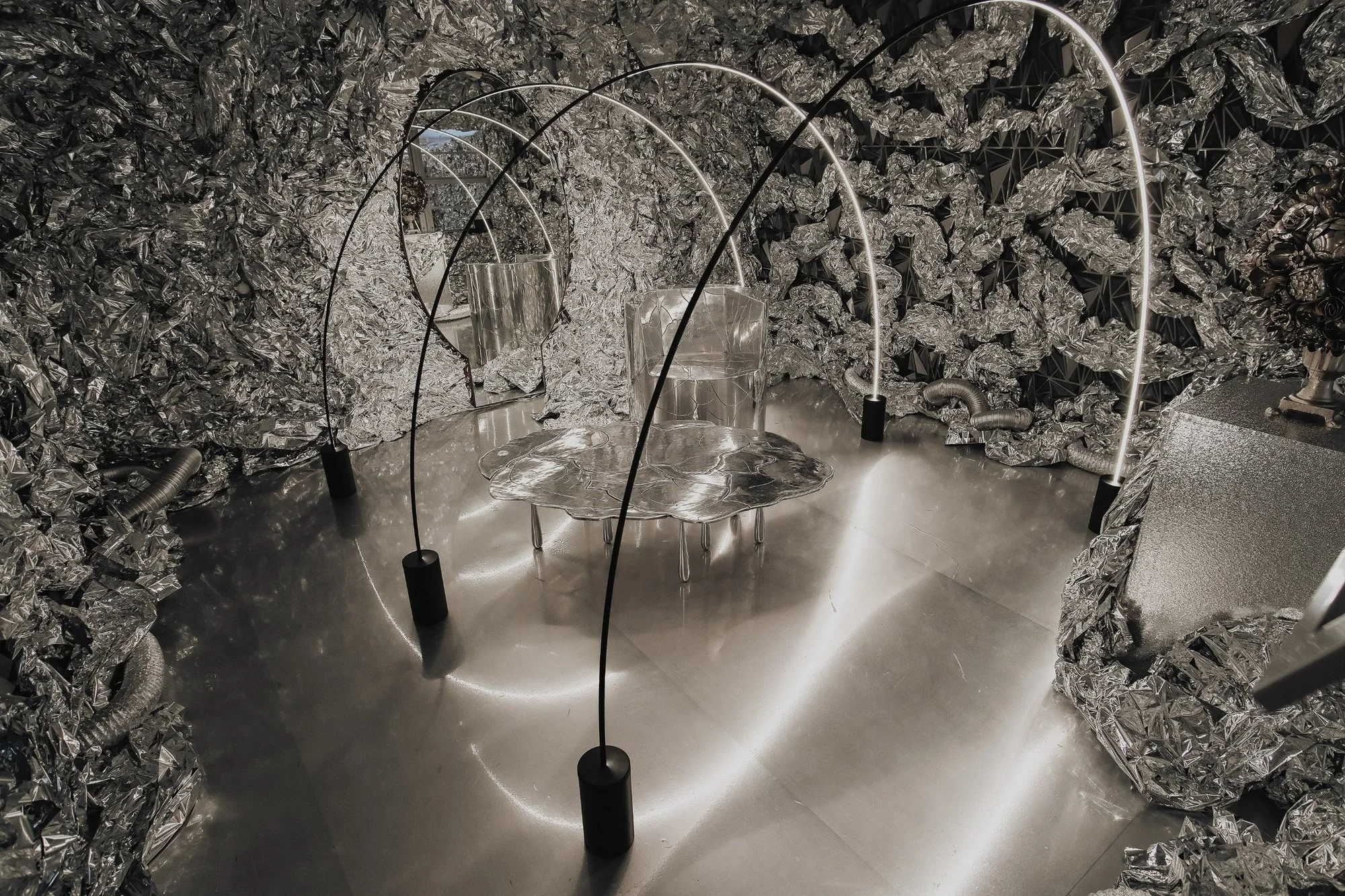 Cosmic diorama: arcs of light over a melted-aluminum table and chair inside a cave of crumpled silver foil
