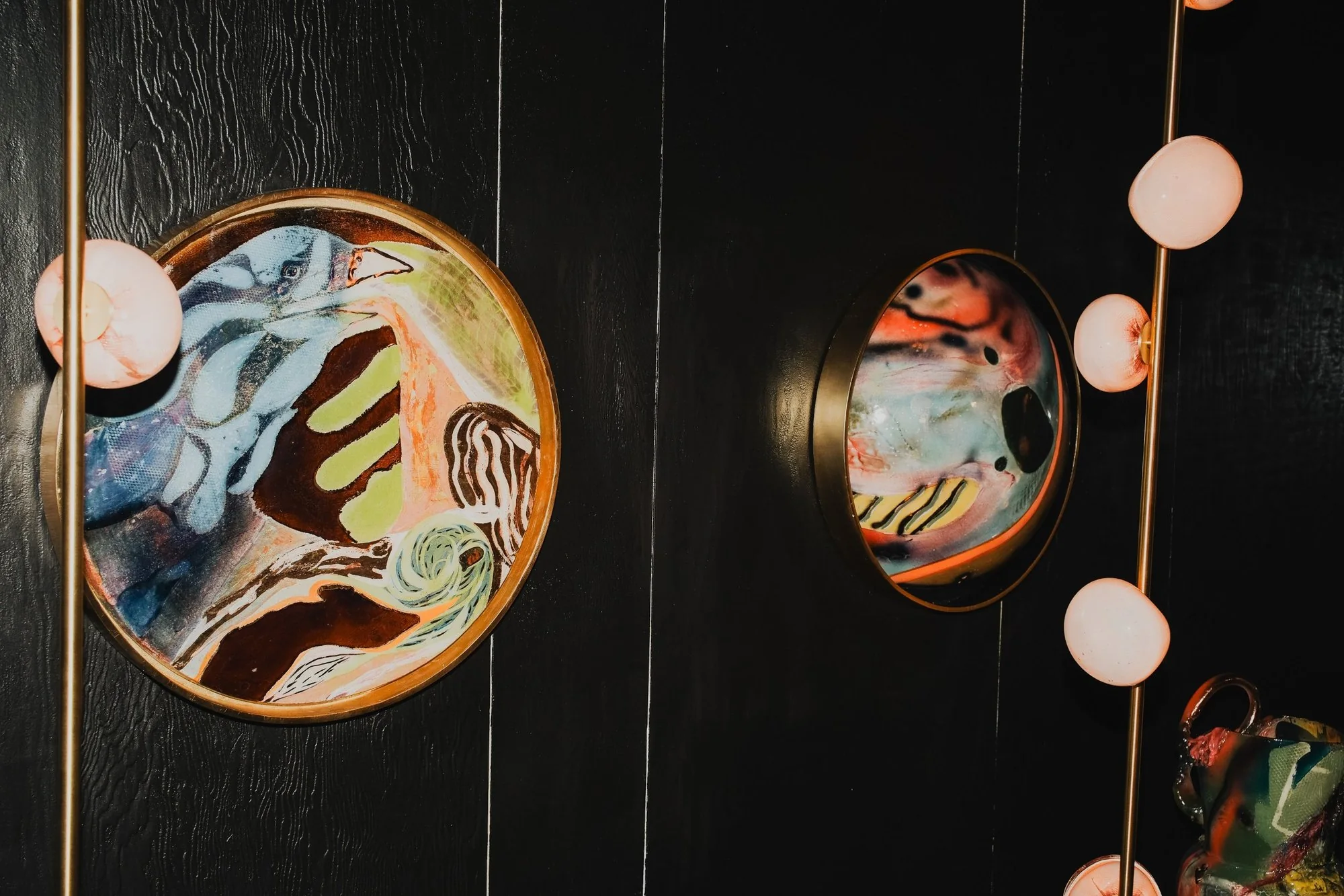 Groovy diorama: round painted artworks in brass frames on a black wood wall, lit by glass globe lights on brass poles